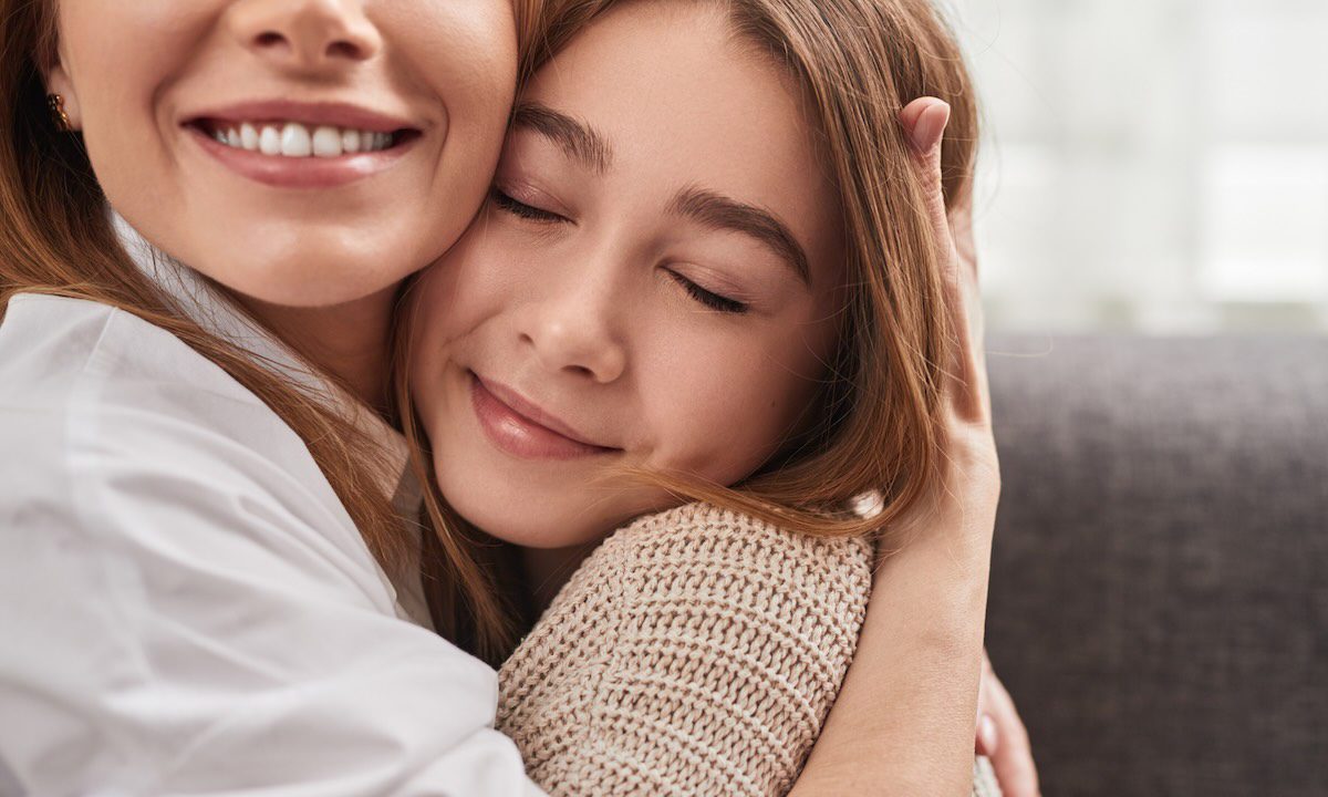 Crop happy loving mom smiling widely while embracing daughter sitting on couch with closed eyes at home