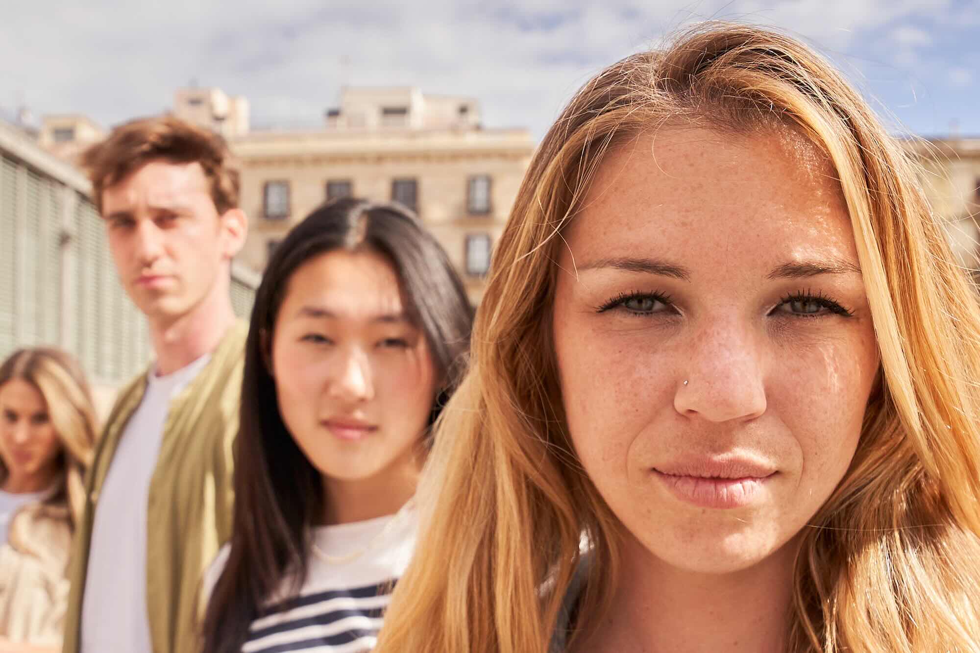 Focus on serious young blonde woman looking at camera on street group multiracial friends in background. Confident people together posing portrait. Colleagues sightseeing on vacation. Generation z.