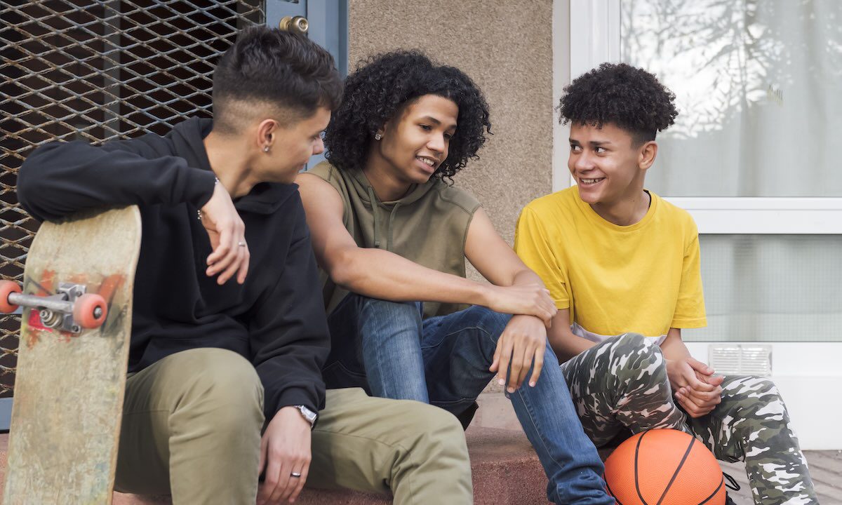 three multiracial male teenagers with basketball and skateboard having fun and talking sitting together on street, concept of youth and urban lifestyle