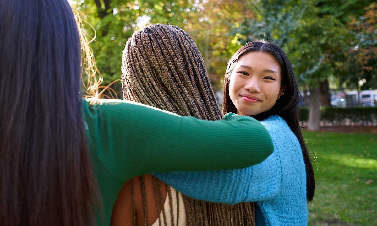 Three Female Friends Embracing At A Public Park, While One Of Them Turns To The Camera Smiling.