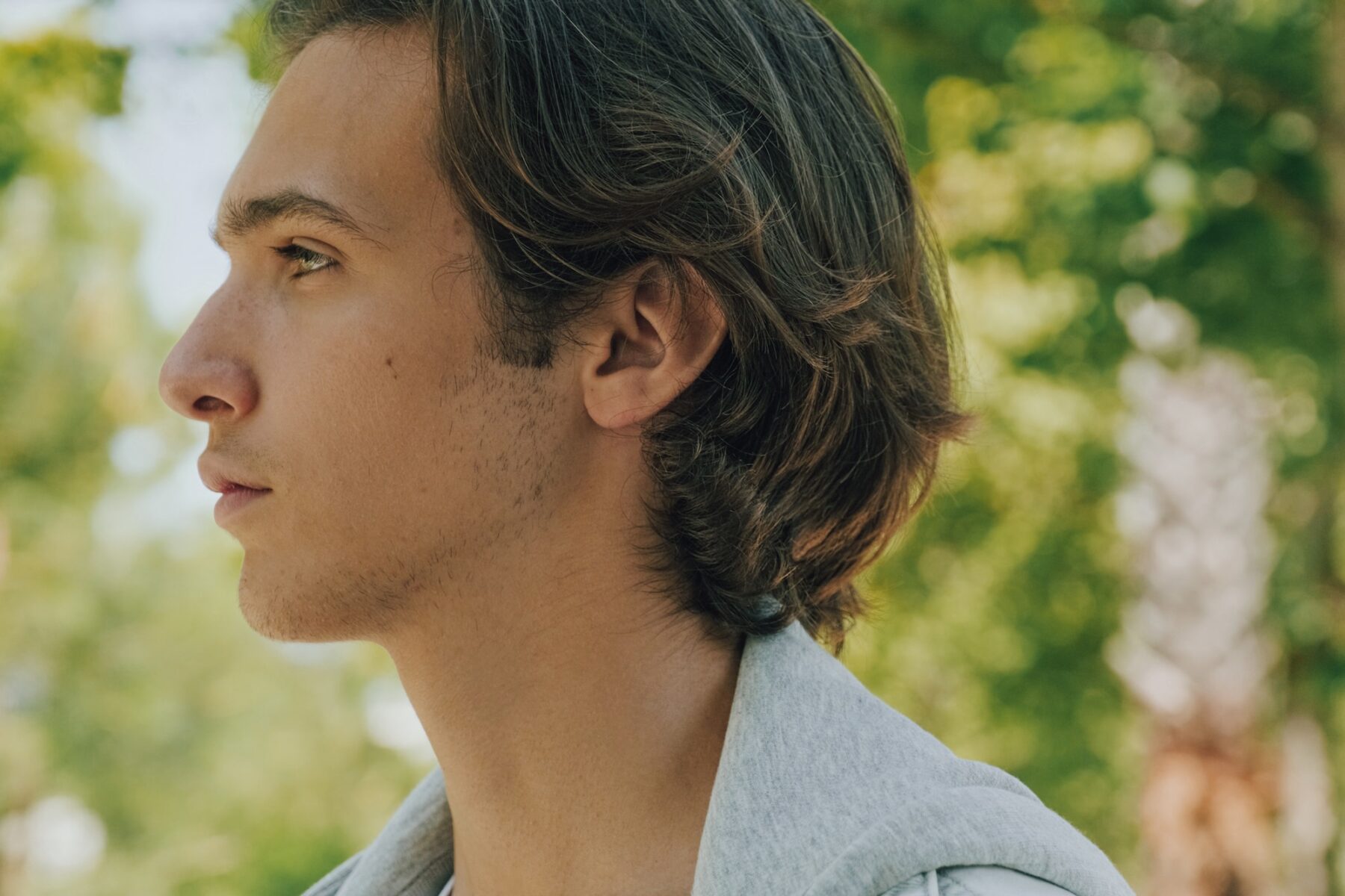 Confident teenager is looking away, outdoor portrait. Handsome face of a male model with brown hair at city. Closeup profile portrait of handsome confident young man at the park in summer.