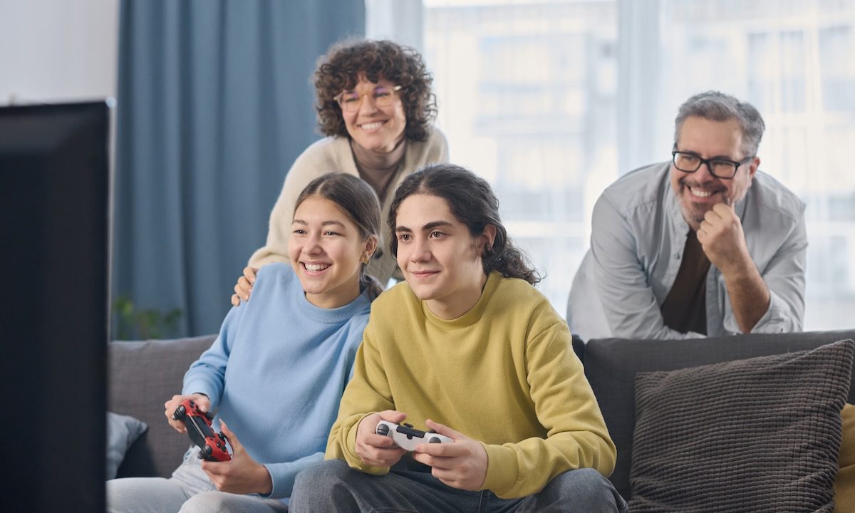 Happy children using joysticks to play video game while parents standing behind them in the room