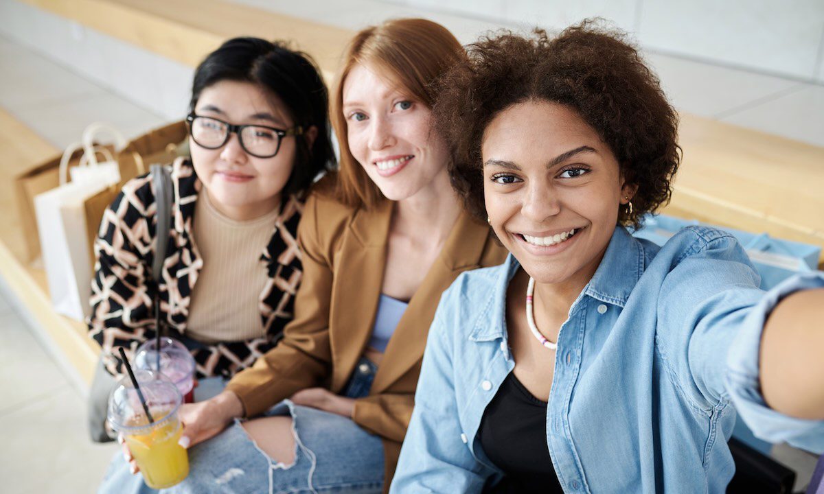 Multiracial group of friends taking selfie together sitting on bench at shopping center