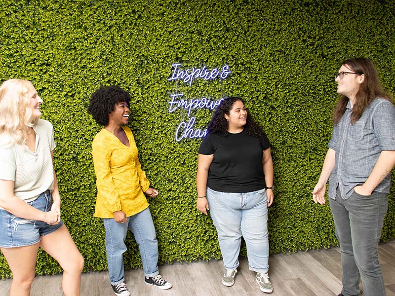 four young adults standing in front of a neon sign reading "inspire and empower change"